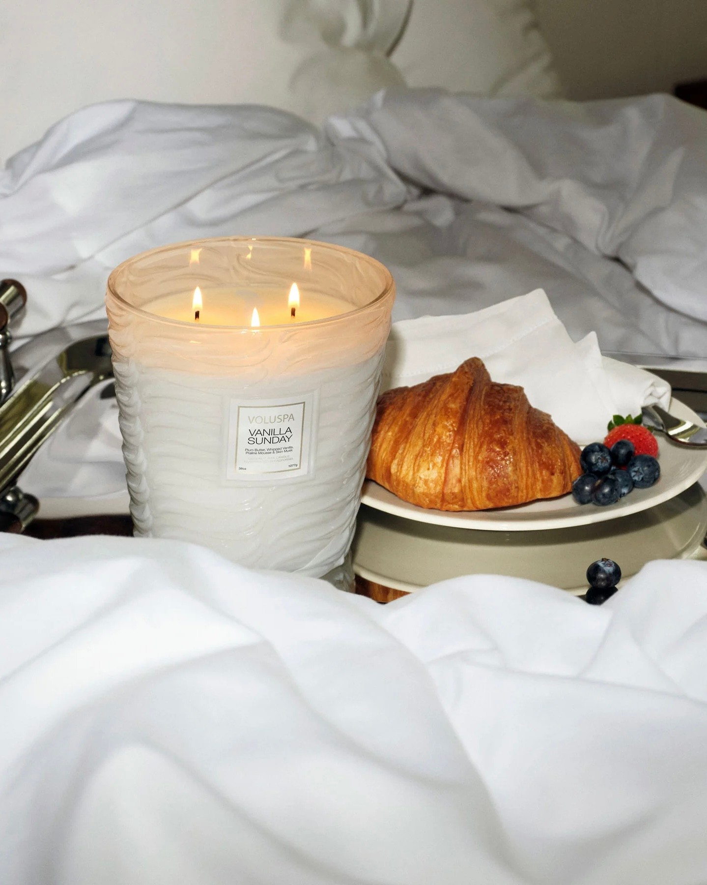 Voluspa candle lit on a tray of breakfast in bed with a croissant and berries